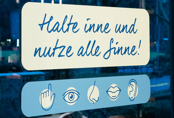 Aufkleber auf der Glasfassade der Hotellobby mit Text: "5 Jahre - 5 Sinne - ein Augenblick. Halte inne und nutze alle Sinne!" Darunter Grafiken von einer Hand, einem Auge, einer Nase, Lippen und einem Ohr.