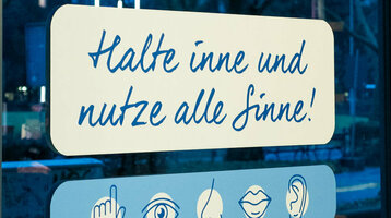 Aufkleber auf der Glasfassade der Hotellobby mit Text: "5 Jahre - 5 Sinne - ein Augenblick. Halte inne und nutze alle Sinne!" Darunter Grafiken von einer Hand, einem Auge, einer Nase, Lippen und einem Ohr.