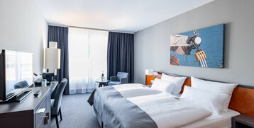 Helles Hotelzimmer mit Doppelbett, modernem Dekor, Schreibtisch, Sessel und großem Fenster im ATLANTIC Hotel Vegesack.