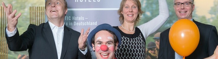 Klinikclown im ATLANTIC Hotel Galopprennbahn Ein fröhlicher Clown mit bunten Luftballons posiert mit drei lächelnden Menschen in einem Hotel.