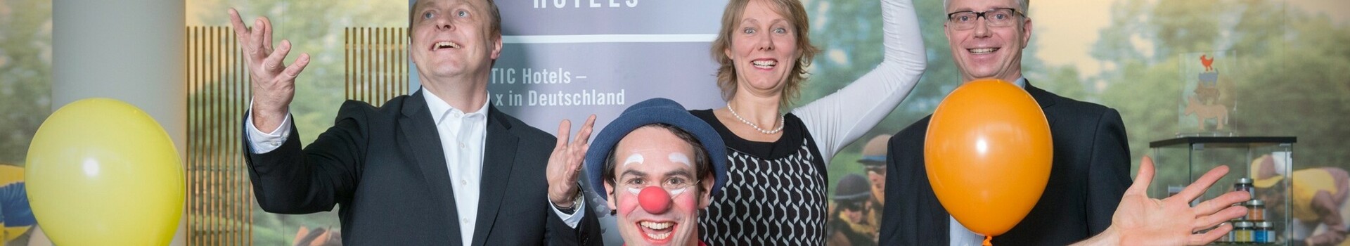 Ein fröhlicher Clown mit bunten Luftballons posiert mit drei lächelnden Menschen in einem Hotel.