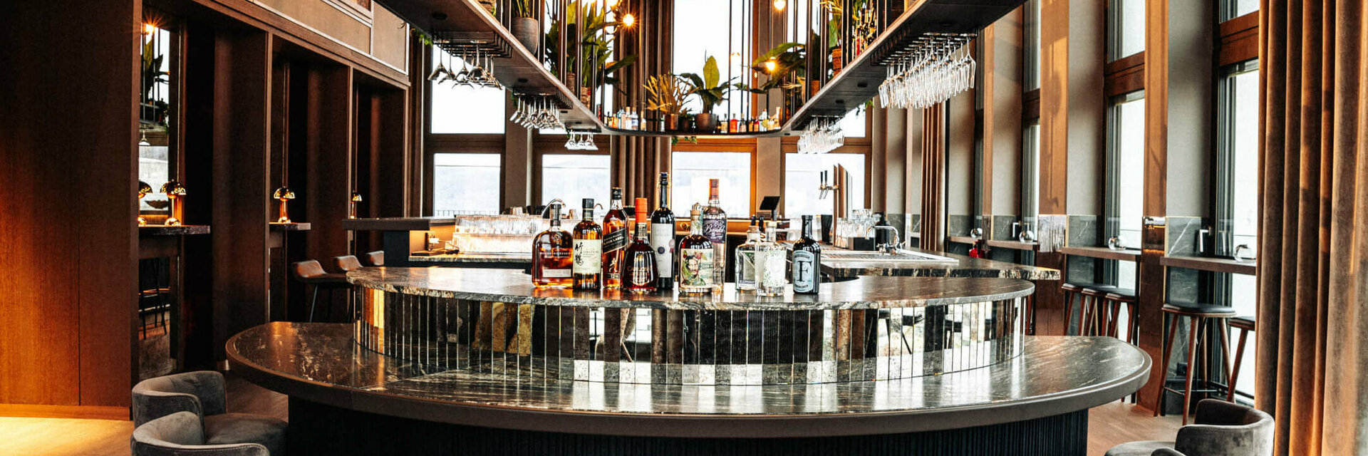 Elegant hotel bar with round counter, bottles, glasses and plants, surrounded by high windows and stylish seating.
