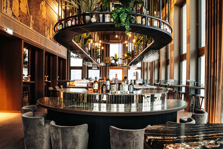 Elegante Hotelbar mit runder Theke, Flaschen, Gläsern und Pflanzen, umgeben von hohen Fenstern und stilvollen Sitzgelegenheiten.
