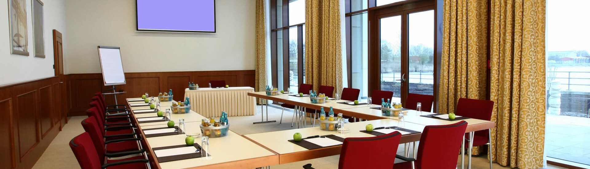 Tagungsraum Travemünde | ATLANTIC Hotel Wilhelmshaven Konferenzraum im ATLANTIC Hotel Wilhelmshaven mit U-förmiger Tischanordnung, roten Stühlen und Blick auf eine Terrasse.