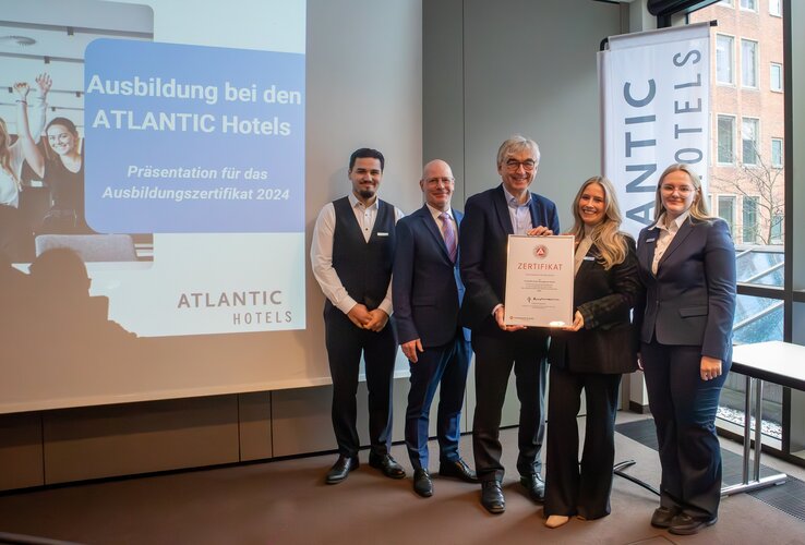 Fünf Personen posieren mit einem Zertifikat vor einer Präsentation über Ausbildung bei ATLANTIC Hotels.
