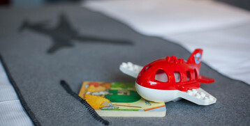 Rotes Spielzeugflugzeug und Buch auf grauer Decke mit Flugzeugsilhouette im ATLANTIC Hotel Airport Bremen.
