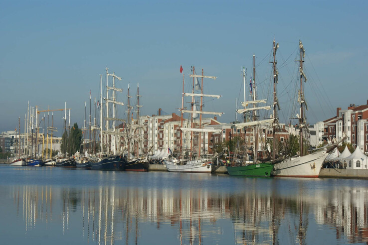 Sailing ships in the harbor, reflected in the calm water, with modern buildings in the background under a clear blue sky.