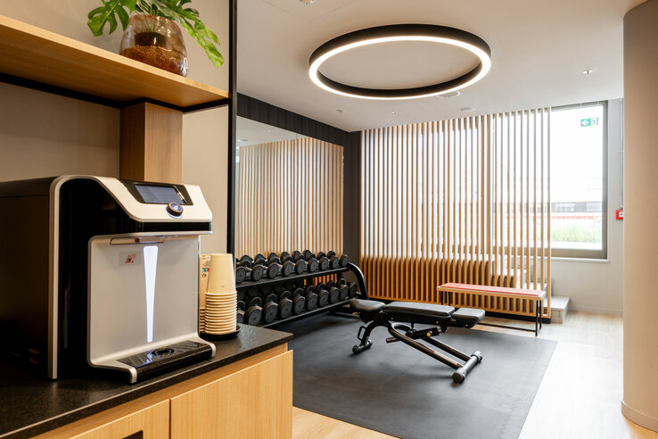 Moderner Fitnessraum im ATLANTIC Hotel Heidelberg mit Hanteln, Trainingsbank und Getränkespender.