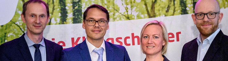 Vier Personen in Anzügen halten Auszeichnungen vor einem Banner mit "energie konsens"-Logo.