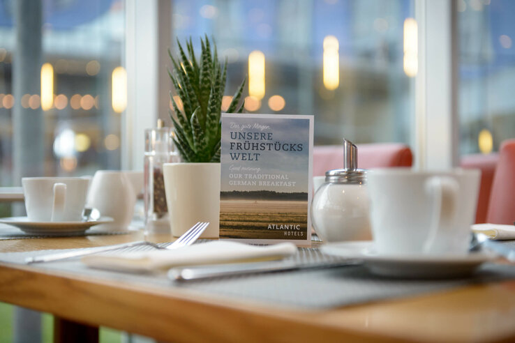 Frühstückstisch im ATLANTIC Hotel mit Kaffeetassen, Pflanze und Werbekarte für traditionelles deutsches Frühstück.