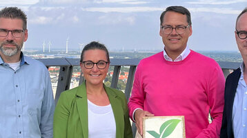 Four people on a viewing platform, one holding a Greensign hotel sign, wind turbines and city view in the background.