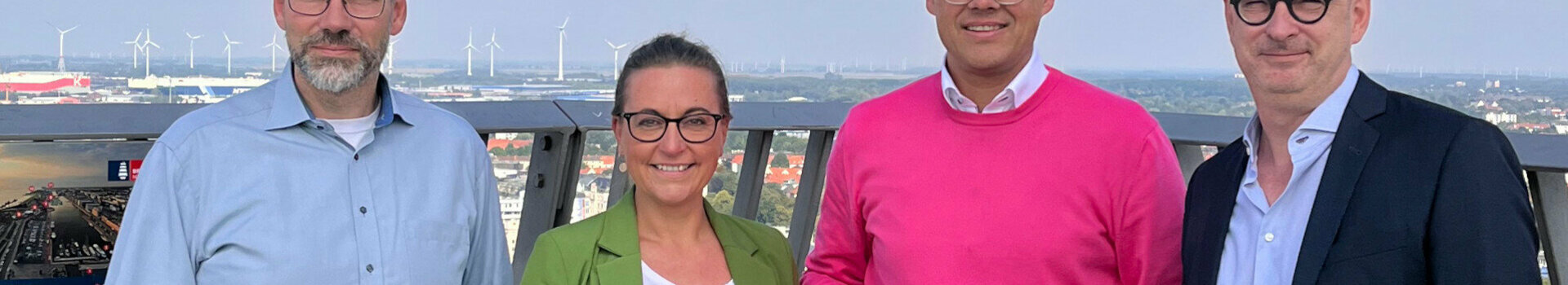 GreenSign Zertifizierung | ATLANTIC Hotels Vier Personen auf einer Aussichtsplattform, eine hält ein Greensign Hotel-Schild, im Hintergrund Windräder und Stadtansicht.