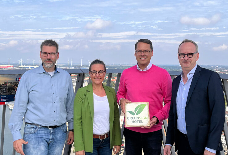 GreenSign Zertifizierung | ATLANTIC Hotels Vier Personen auf einer Aussichtsplattform, eine hält ein Greensign Hotel-Schild, im Hintergrund Windräder und Stadtansicht.