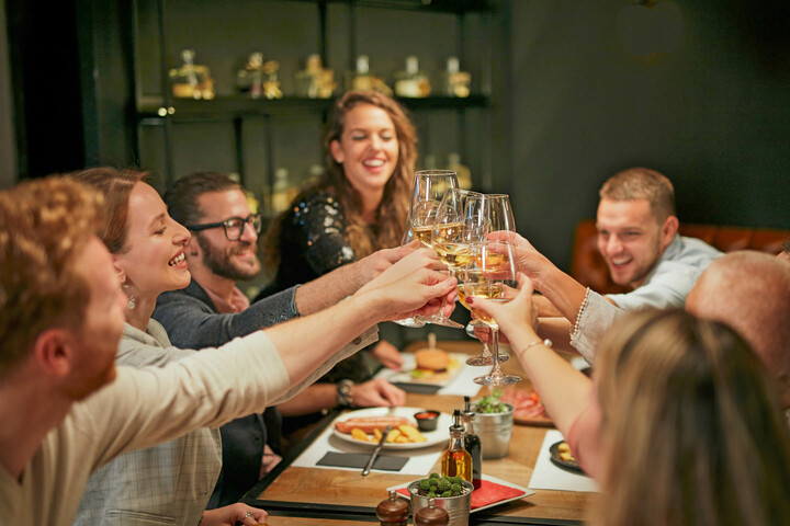 ATLANTIC Hotel Münster Speisen & Getränke Eine Gruppe von Menschen stößt fröhlich mit Weingläsern an einem gedeckten Tisch an.