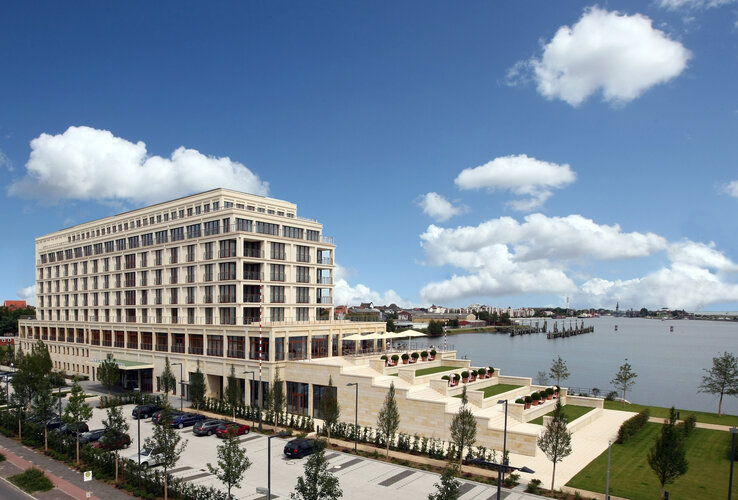 Modernes Hotel am Wasser mit großen Fenstern, Terrassen und gepflegtem Garten, umgeben von blauem Himmel und Wolken.