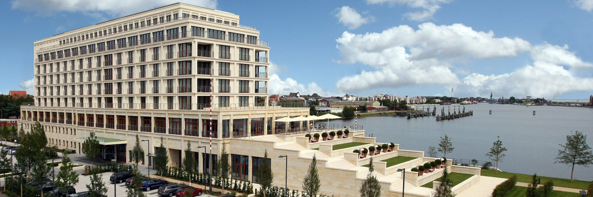 Modern hotel on the waterfront with large windows, terraces and well-tended garden, surrounded by blue sky and clouds.
