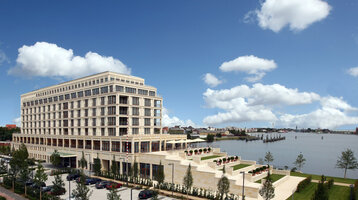 Modernes Hotel am Wasser mit großen Fenstern, Terrassen und gepflegtem Garten, umgeben von blauem Himmel und Wolken.