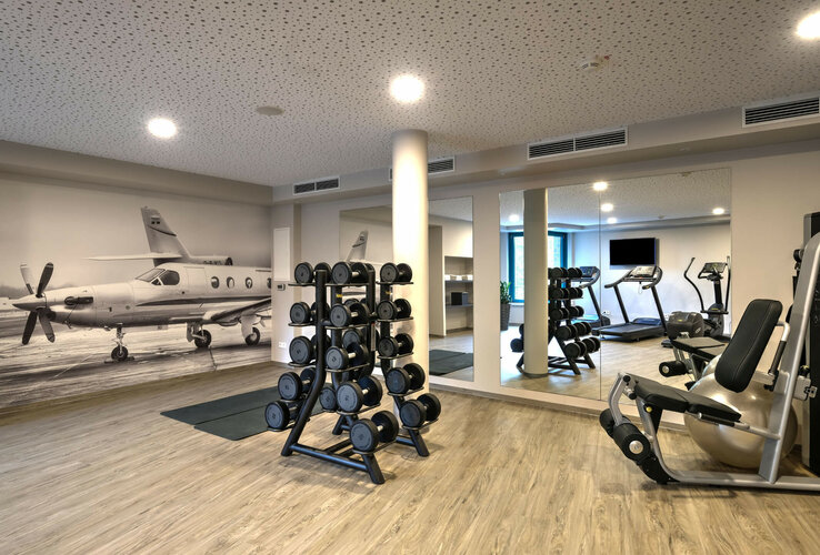 Fitness room at the ATLANTIC Hotel Airport Bremen with weights, fitness equipment and a picture of an airplane on the wall.