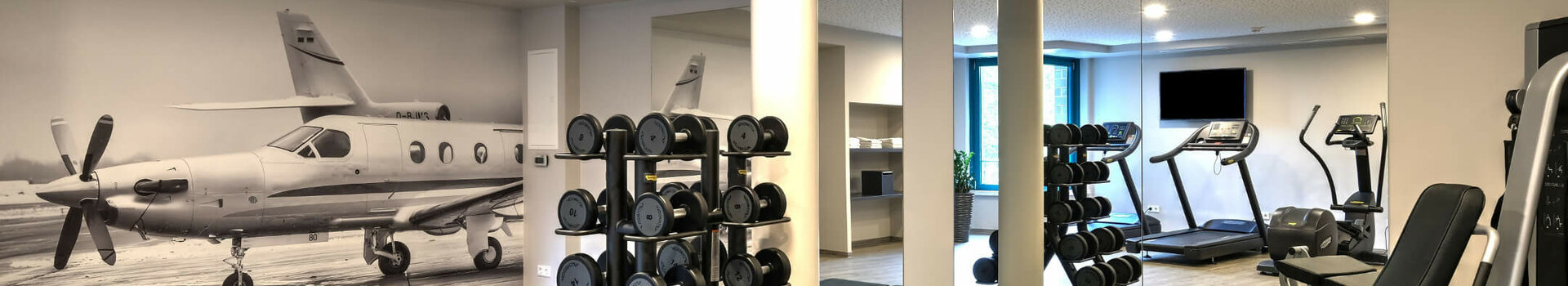 Fitness area at ATLANTIC Hotel Airport Bremen Fitness room at the ATLANTIC Hotel Airport Bremen with weights, fitness equipment and a picture of an airplane on the wall.