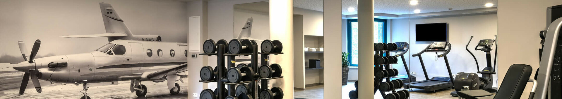Fitness area at ATLANTIC Hotel Airport Bremen Fitness room at the ATLANTIC Hotel Airport Bremen with weights, fitness equipment and a picture of an airplane on the wall.