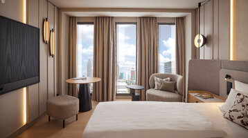 Modern eingerichtetes Hotelzimmer im ATLANTIC Hotel Frankfurt mit großem Bett, Sitzbereich mit Sessel und kleinem Tisch sowie bodentiefen Fenstern, durch die eine Stadtansicht mit Hochhäusern zu sehen ist.