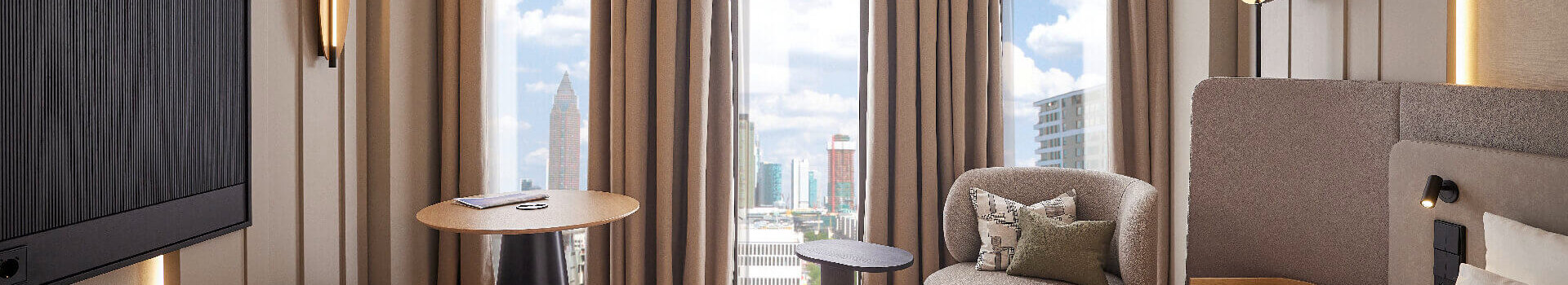 Modern eingerichtetes Hotelzimmer im ATLANTIC Hotel Frankfurt mit großem Bett, Sitzbereich mit Sessel und kleinem Tisch sowie bodentiefen Fenstern, durch die eine Stadtansicht mit Hochhäusern zu sehen ist.