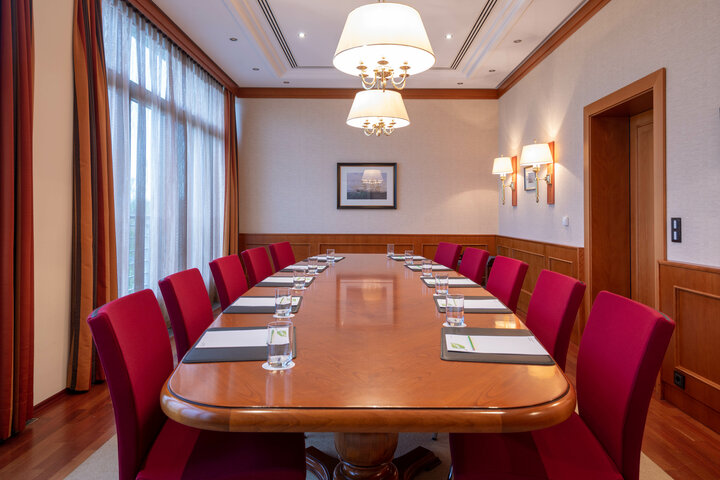 Konferenzraum mit langem Holztisch, roten Stühlen, Notizblöcken und Wassergläsern, große Fenster mit Vorhängen.