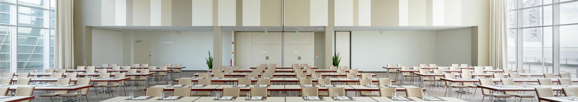 Conference room Conference 1 at the ATLANTIC Hotel Sail City in Bremerhaven Modern conference room with rows of tables and chairs, large windows overlooking the water.