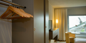 Modernes Hotelzimmer mit Kleiderschrank, Flachbild-TV, Sessel, Tisch und großem Fenster mit Blick nach draußen.