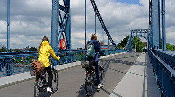 Fahrrad fahren in Wilhelmshaven und Umgebung | Leihfahrräder vom ATLANTIC Hotel Wilhelmshaven Zwei Personen radeln über eine blaue Stahlbrücke unter bewölktem Himmel.