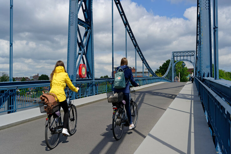 Zwei Personen radeln über eine blaue Stahlbrücke unter bewölktem Himmel.