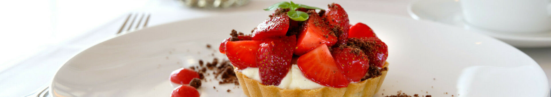 Atlantic Hotels Newsletter Restaurant Erdbeertörtchen mit Sahne und Schokostreuseln auf weißem Teller, dekoriert mit Erdbeersauce.
