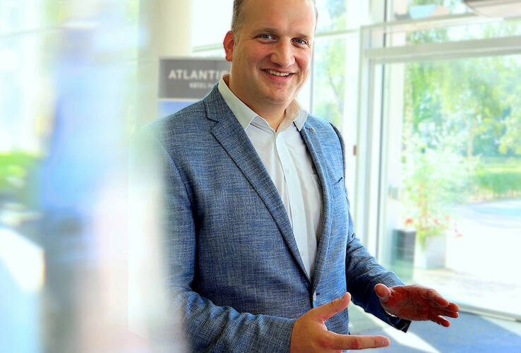 A smiling man in a blue suit stands in a bright, modern hotel entrance area.