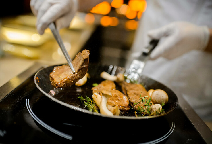 Koch brät Fleisch und Pilze in einer Pfanne, mit Kräutern garniert. Hände mit Handschuhen bedienen die Utensilien.