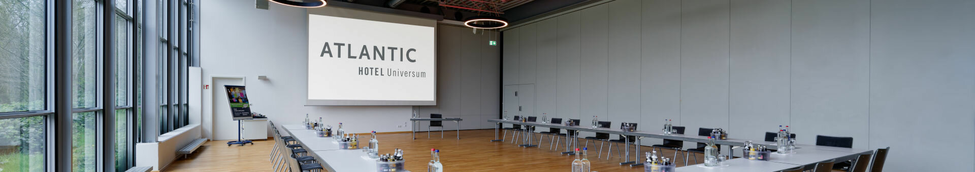 Moderner Konferenzraum im ATLANTIC Hotel Universum mit U-förmiger Tischanordnung, großen Fenstern und Präsentationsleinwand.