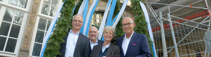 Vier Personen stehen lächelnd unter einem dekorativen Kranz mit blauen und weißen Bändern vor einem historischen Gebäude.