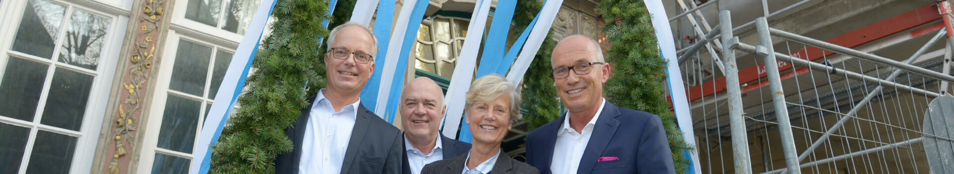 Vier Personen stehen lächelnd unter einem dekorativen Kranz mit blauen und weißen Bändern vor einem historischen Gebäude.