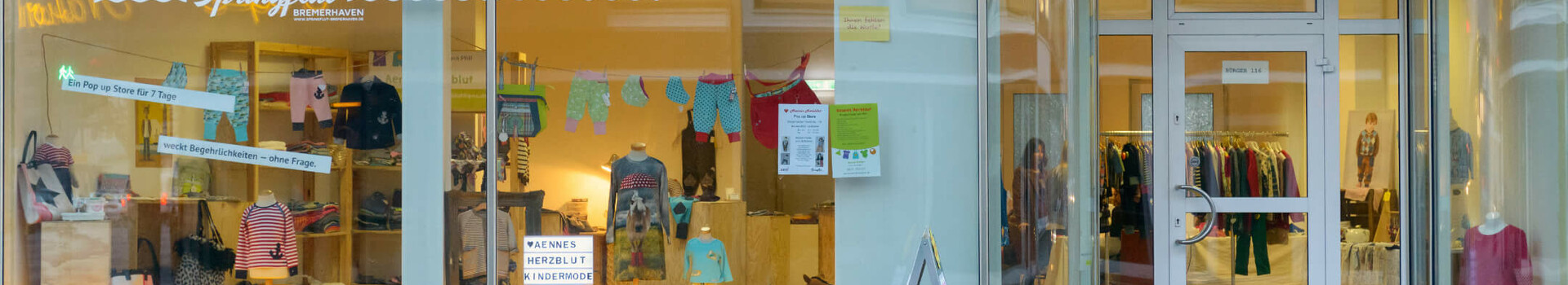 ATLANTIC Hotel Sail City | Upcycling-Offensive "GRÜNER FADEN" Schaufenster eines Kindermodegeschäfts mit bunter Kleidung und Dekoration, daneben Glastür zum Ladeninneren.