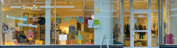 Schaufenster eines Kindermodegeschäfts mit bunter Kleidung und Dekoration, daneben Glastür zum Ladeninneren.