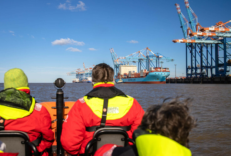 Personen in roten Jacken auf einem Boot, Blick auf Containerhafen und Kräne in Bremerhaven bei blauem Himmel.