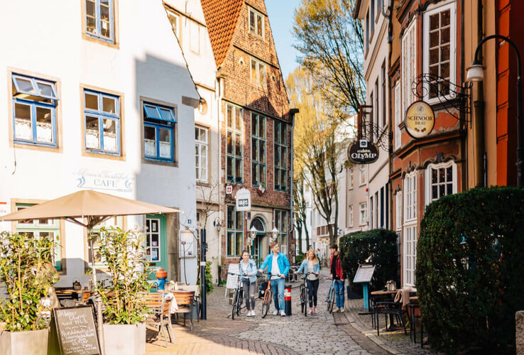 Pittoreske Altstadtgasse mit Fachwerkhäusern, Café und Menschen mit Fahrrädern bei sonnigem Wetter.