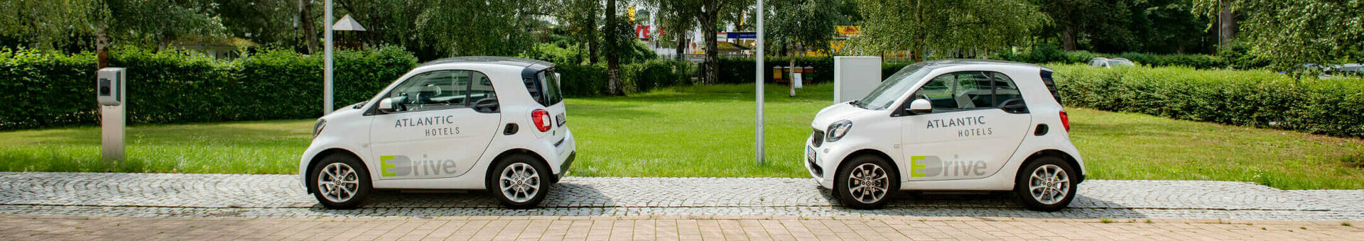 Electric filling stations | Service of the ATLANTIC Hotels Two white electric Smarts with the "Atlantic Hotels" logo are parked next to each other on a paved path, surrounded by trees.