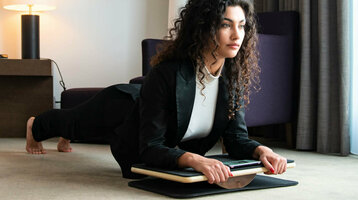 Woman in business attire doing plank exercise on balance board in modern hotel room.