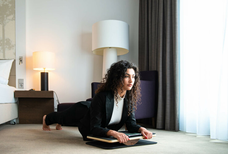 Woman in business attire doing plank exercise on balance board in modern hotel room.