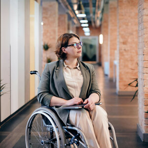 Person im Rollstuhl in einem modernen, hellen Korridor mit Pflanzen und Backsteinwänden.