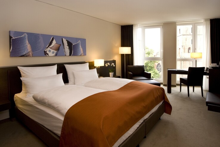 Modernes Hotelzimmer mit Doppelbett, orangefarbener Decke, Schreibtisch, Sesseln und großem Fenster mit Blick auf Kirche.