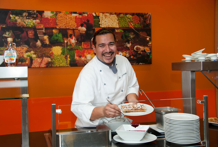 Koch im ATLANTIC Hotel Kiel serviert lächelnd frische Waffeln vor einem farbenfrohen Marktbild.