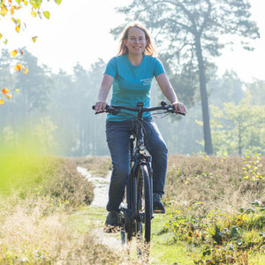 Frau auf einem Fahrrad fährt auf einem schmalen Pfad durch eine sonnige, bewaldete Landschaft.