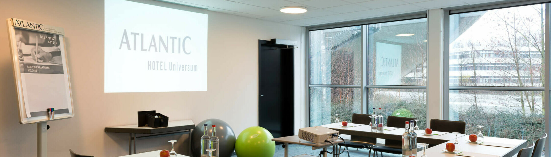 ATLANTIC Hotel Universum – Saal 2 Heller Konferenzraum im ATLANTIC Hotel Universum mit Tischen, Stühlen, Projektor und Flipchart, Blick nach draußen.