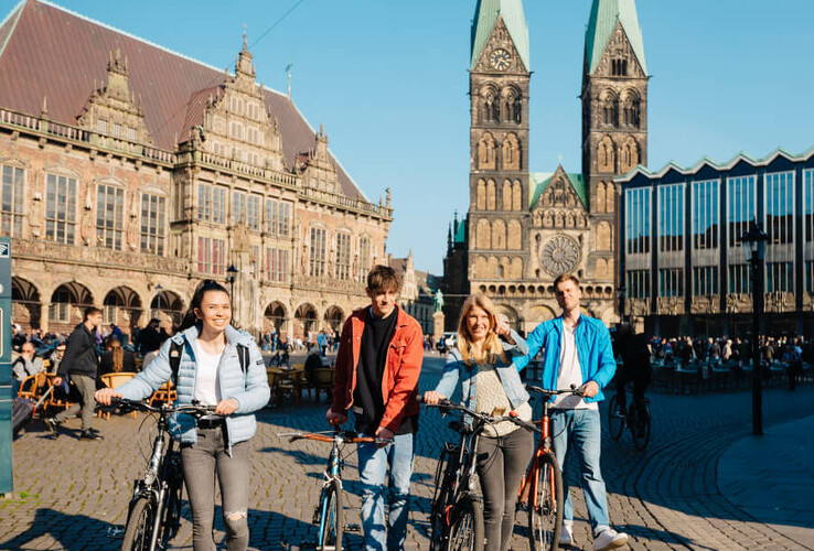 Vier Personen mit Fahrrädern vor historischer Architektur und zwei Kirchtürmen auf einem sonnigen Platz.